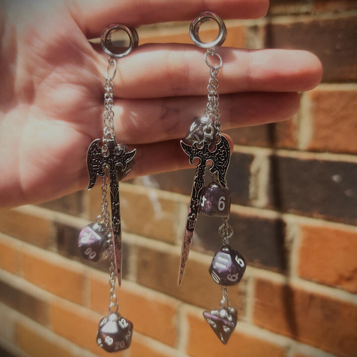 A pair of silver 00g tunnels with halberds juxtaposed with purple dice being held up against a brick wall.